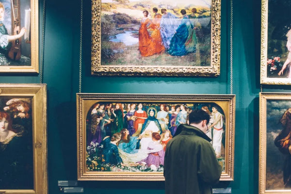A visitor in a dark coat observing a collection of framed classical paintings in an art gallery with deep green walls.