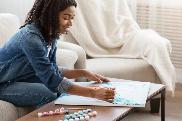 A smiling woman painting a canvas using a paint-by-numbers kit while sitting on a cozy couch, with paint pots arranged on a coffee table.