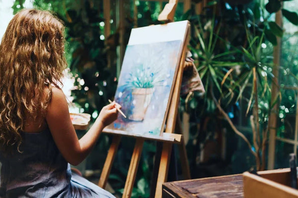 A woman with curly hair painting on a canvas in a bright, plant-filled room. She is using an easel and appears focused on her artwork.