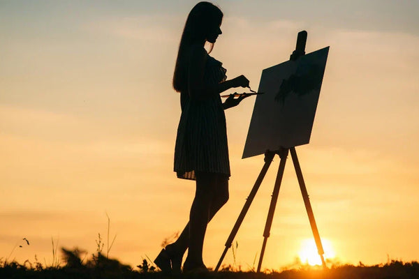 A silhouette of a woman painting on a canvas outdoors during sunset, standing in front of an easel with a brush in hand.