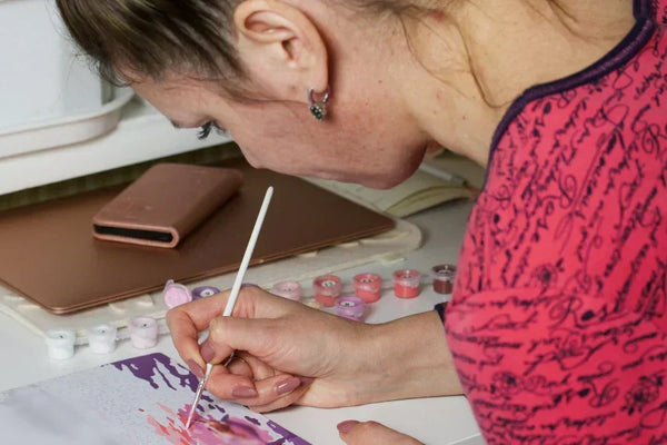 A woman concentrating while painting a paint-by-numbers canvas at a desk, surrounded by small paint pots and art supplies.