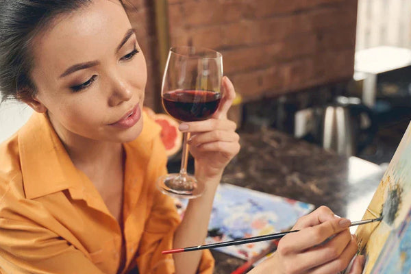 A woman enjoying a relaxing painting session while holding a glass of red wine, working on a canvas in a cozy indoor setting.