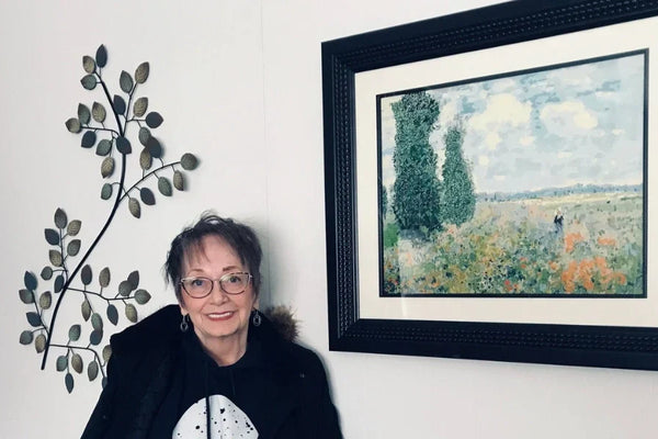 A smiling woman standing next to a framed impressionist-style painting on a wall, showcasing her completed paint-by-numbers artwork.