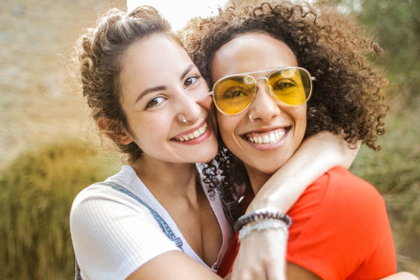 Two smiling women embracing outdoors, one wearing yellow sunglasses and the other in a white top, capturing a moment of friendship and happiness.
