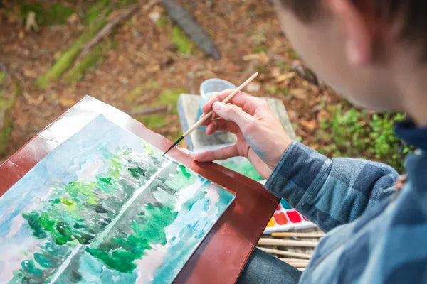 A person painting a watercolor landscape outdoors, carefully using a fine brush to add details to a nature-inspired scene.