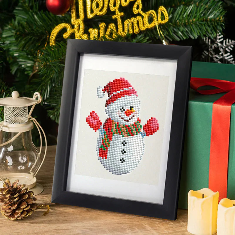 Framed mini diamond painting of a festive snowman wearing a Santa hat and scarf, displayed in a cozy Christmas setting with gifts, candles, and holiday décor.