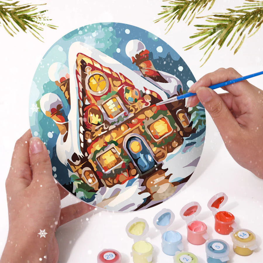 Festive round paint by numbers placemat featuring a colorful gingerbread house, with a hand painting the details using a brush and acrylic paints. A perfect holiday DIY craft.